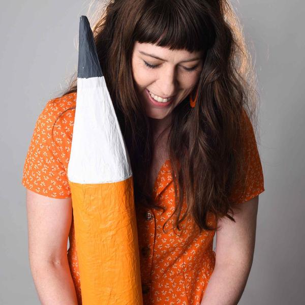 A woman with brown hair wearing an orange dress and holding a large fake pencil.